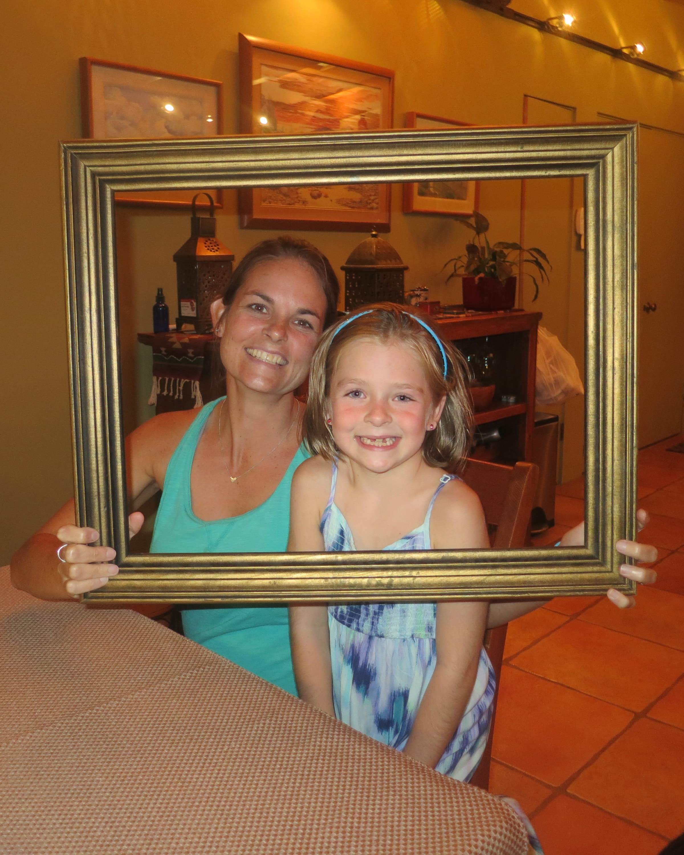 Woman in a teal tank top and young girl in a blue sundress and headband smiling together through a gold picture frame indoors with Southwest artwork on the walls behind them