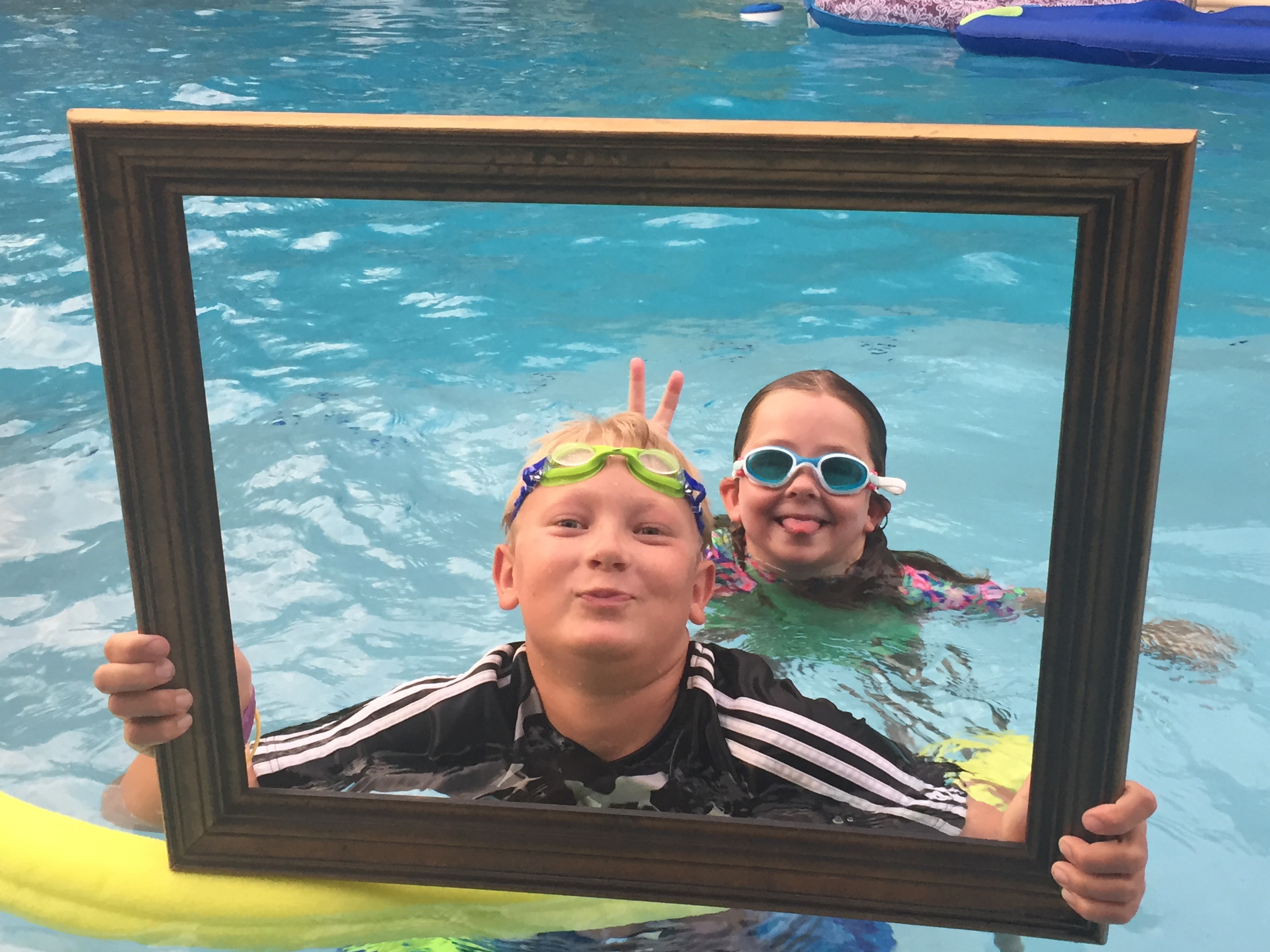 Two kids in swim goggles grinning through a dark wood picture frame in a backyard swimming pool, one giving bunny ears