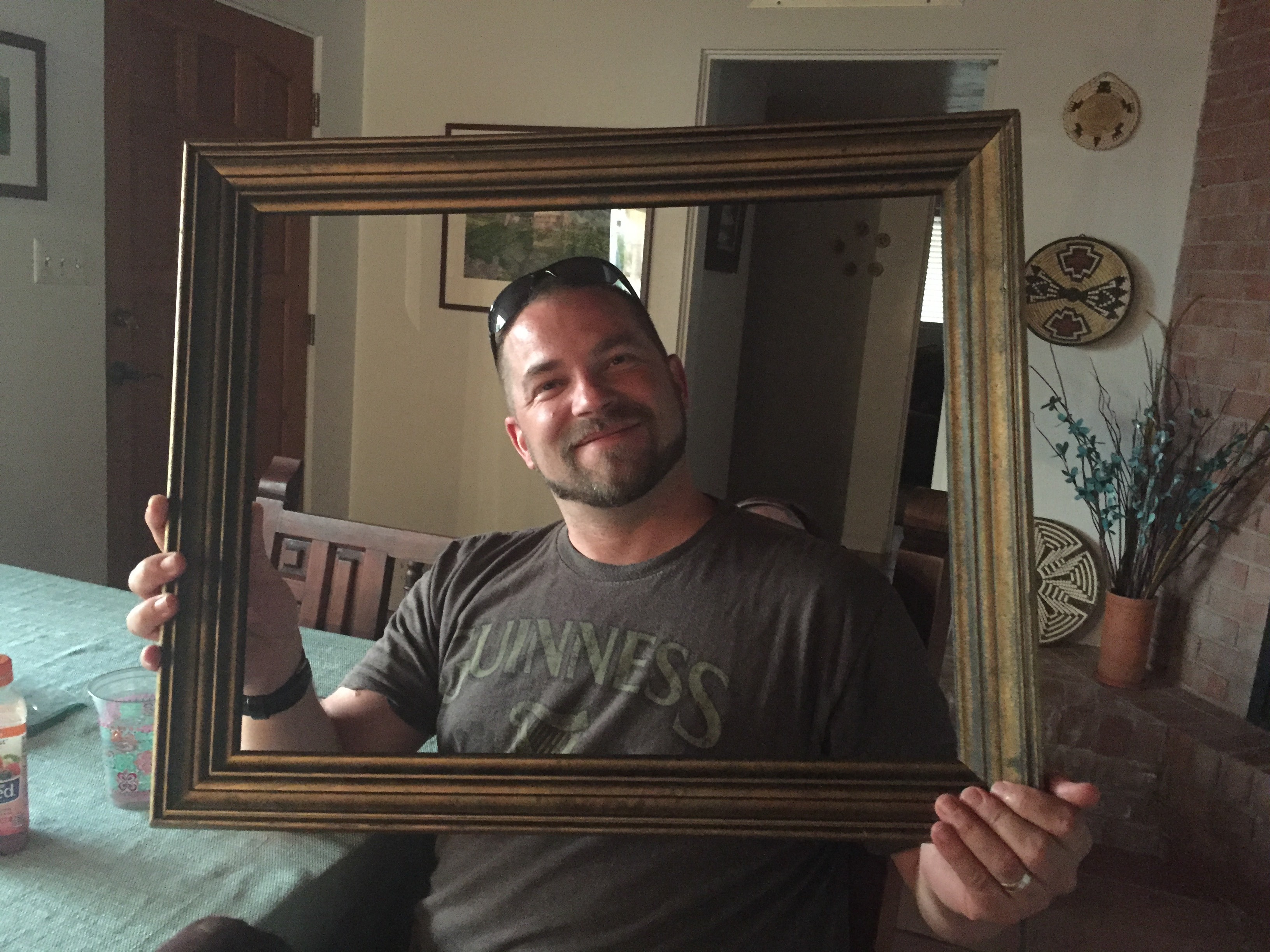 Smiling man in a Guinness shirt holding a large gold picture frame at a dining table with Southwest basket art on the wall behind him