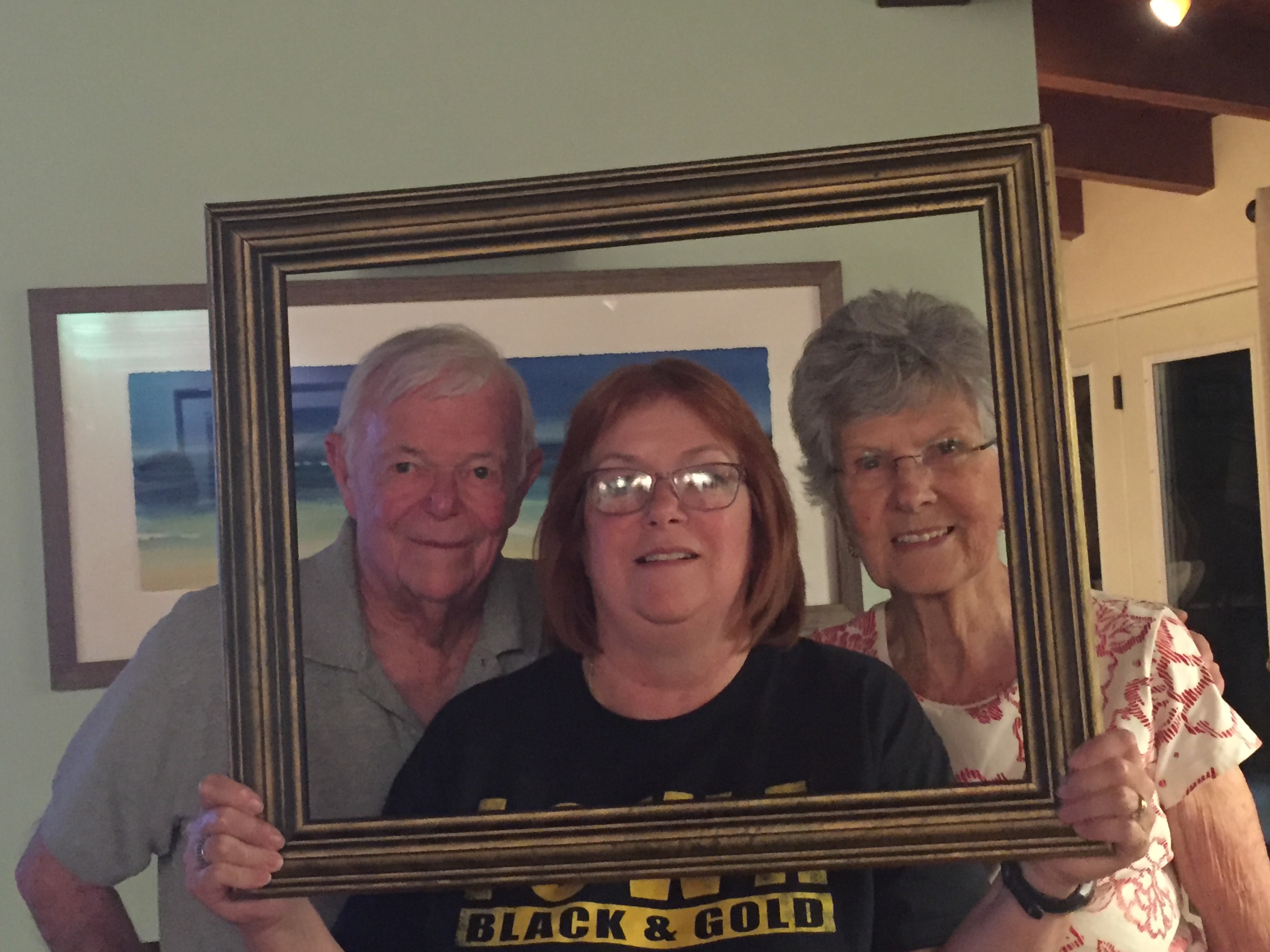 Older gentleman, red-haired woman in an Iowa Black and Gold shirt, and silver-haired woman smiling together through a gold picture frame indoors with a beach painting behind them