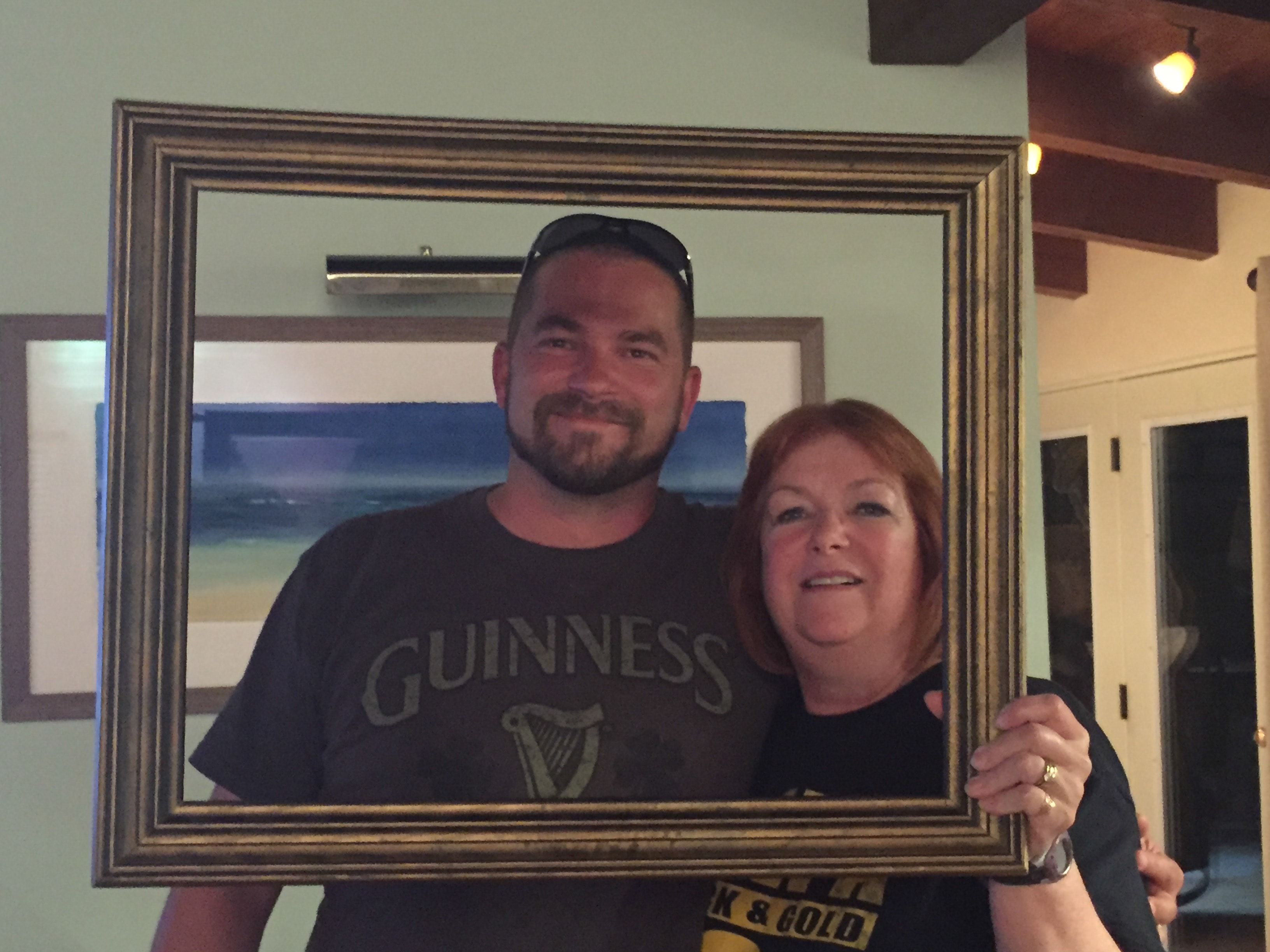 Man in a Guinness shirt and red-haired woman in an Iowa shirt smiling together through a gold picture frame with a beach painting on the wall behind them