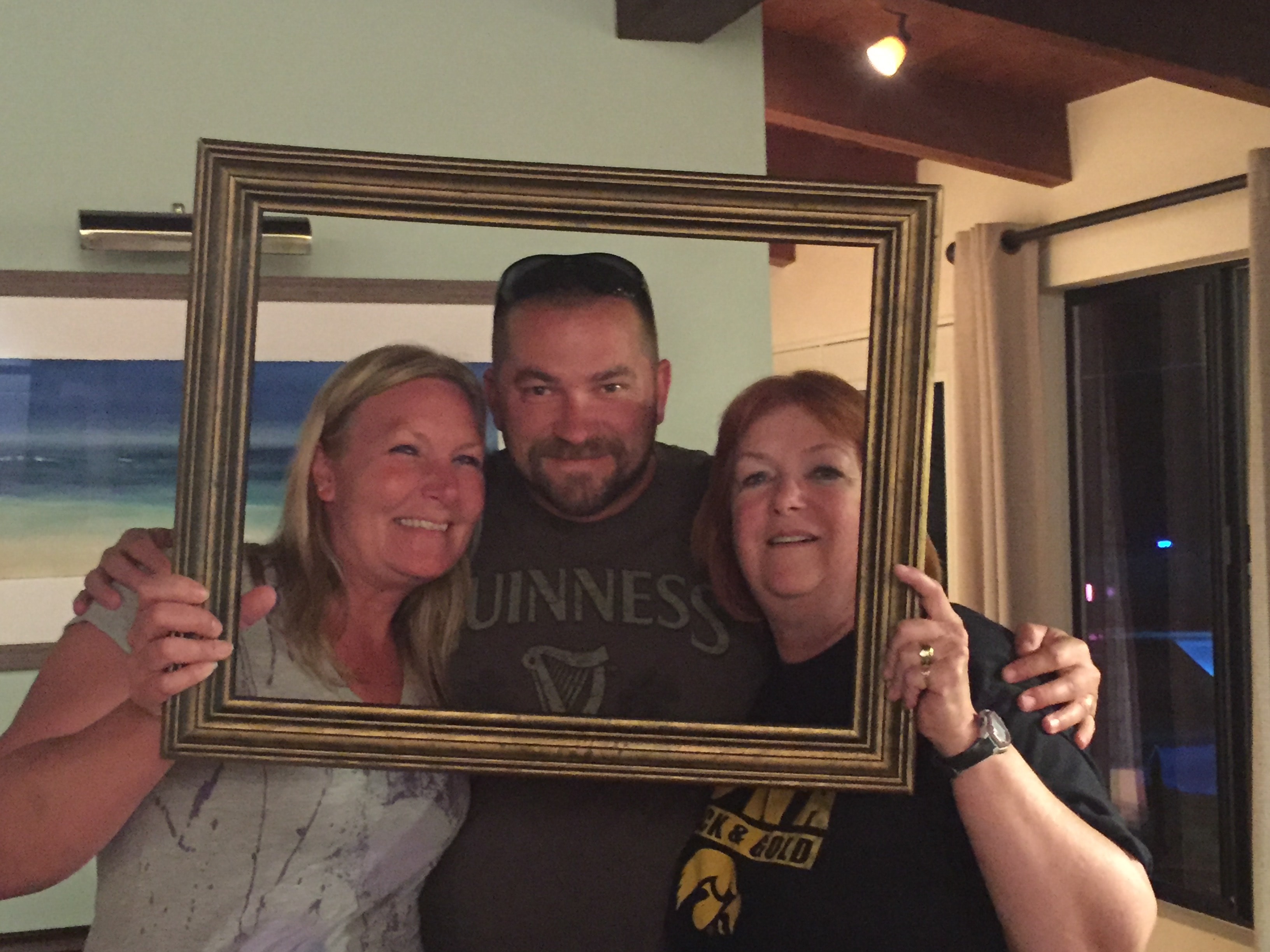 Blonde woman, man in a Guinness shirt, and red-haired woman in an Iowa shirt laughing together through a gold picture frame indoors at night