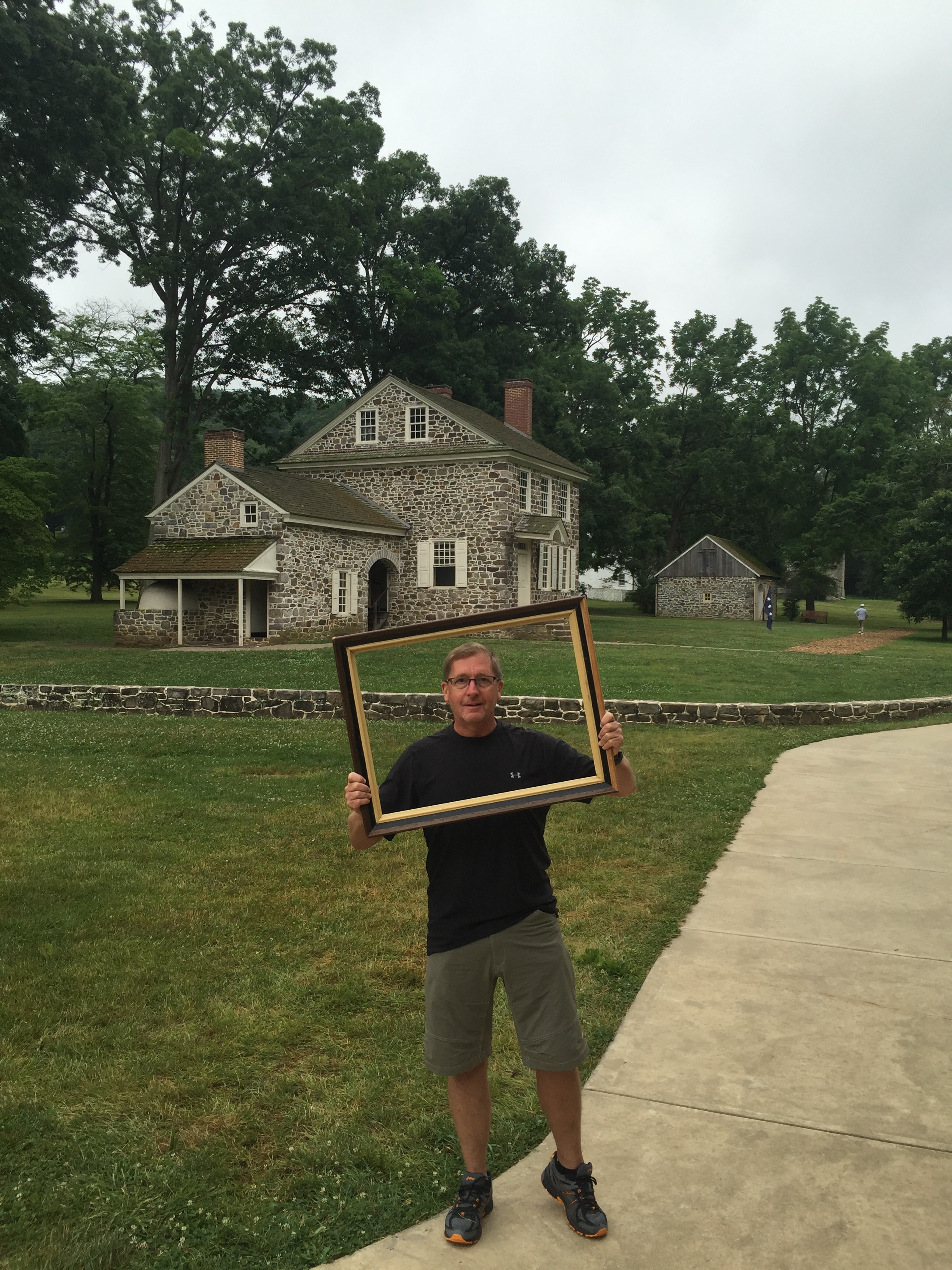 Man in a black Under Armour shirt and olive shorts holding a dark wood and gold picture frame on a sweeping green lawn in front of a two-story historic stone colonial house